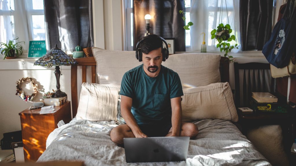A man wearing headphones and using his laptop on his bed